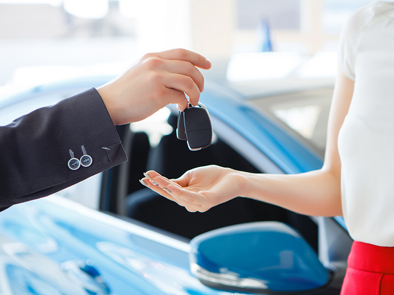 Car salesman handing new car keys to a woman wearing a white top and red pants